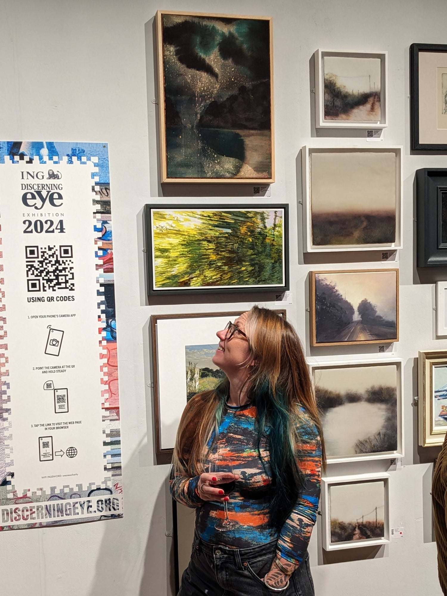 Woman looking at framed artworks on a wall with a calendar titled 'Discerning Eye 2024' to her left.